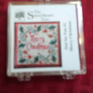 Pode incluir: Uma caixa quadrada de plástico transparente contendo um kit de ponto cruz. O encarte apresenta um fundo branco com uma borda vermelha, um design de guirlanda de azevinho e o texto Merry Christmas. O logotipo The Sweetheart Tree está no canto superior esquerdo.