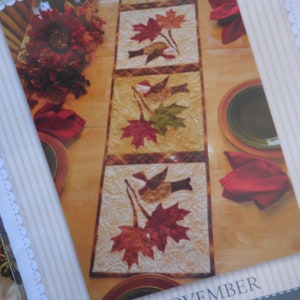 Puede incluir: Un camino de mesa acolchado con un tema otoñal que presenta pájaros y hojas de arce en tonos de rojo, naranja, amarillo y verde. El camino de mesa está hecho por Shabby Fabrics y se titula "Noviembre".