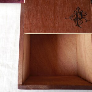 Puede incluir: Una caja de madera con tapa abatible. La tapa tiene un diseño decorativo con un monograma en el centro. La caja está vacía.