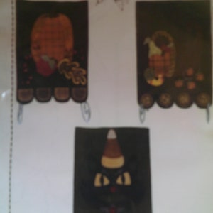 May include: Three felt wall hangings with fall themes. The first features a pumpkin, the second a cornucopia, and the third a black cat with candy corn.