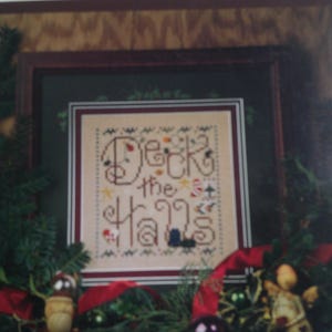 May include: A framed cross-stitch sampler with the words "Deck the Halls" in a decorative font. The sampler is surrounded by a Christmas wreath with red ribbon, ornaments, and greenery. The frame is dark brown.