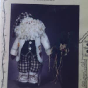 May include: A handcrafted Santa Claus doll, approximately 25 inches tall, with a white beard and a plaid hat and pants. He wears a white coat with black buttons and holds a small bundle of greenery. The doll is a festive holiday decoration.