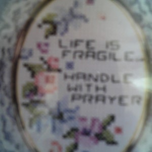 以下が含まれることがあります： 白い楕円形の刺繍枠に、花柄のデザインと「Life is Fragile Handle With Prayer」というテキストが刺繍されています。