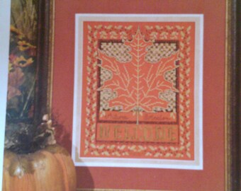 May include: A framed cross-stitch design featuring an orange maple leaf with the words "Autumn Welcome" in a gold font. The design is surrounded by a decorative border of gold leaves.