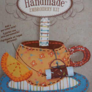 May include: Handmade embroidery kit featuring a colorful teacup with a floral pattern and a small orange slice. The kit includes instructions for adding a personal note.