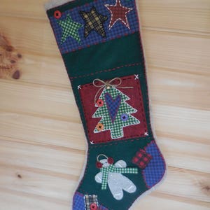 May include: A green Christmas stocking with a patchwork design. The stocking features a red and green plaid star, a green and white plaid star, a brown and white plaid star, a green and white plaid Christmas tree, a red heart, and a gingerbread person.