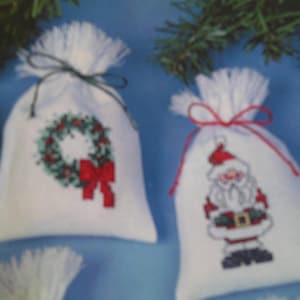 Puede incluir: Dos bolsas de regalo de tela blanca con diseños bordados. Una bolsa presenta una corona verde con un lazo rojo, y la otra bolsa presenta un Papá Noel rojo y blanco.