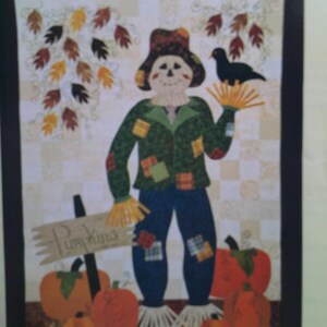 May include: A quilted wall hanging featuring a scarecrow with a crow perched on its hand. The scarecrow wears a green jacket, blue pants, and a brown hat. Pumpkins and autumn leaves decorate the scene. The sign reads "Pumpkins".