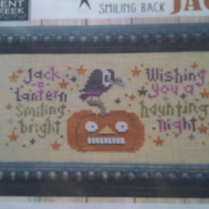 Może przedstawiać: Haft krzyżykowy w ciemnej drewnianej ramie. Projekt przedstawia dynię z krukiem, otoczoną tekstem "Jack Lantern Smiling Bright" i "Wishing You a Haunting Night."
