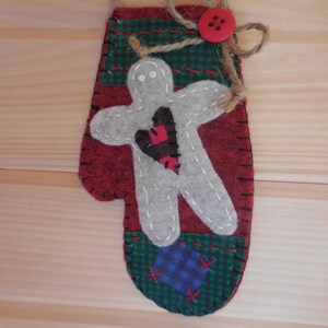 May include: A red felt mitten ornament with a white gingerbread person holding a red heart. The mitten has a green and blue plaid fabric at the bottom and a brown twine bow with a red button.