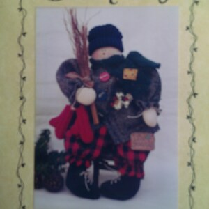 May include: A folk art doll wearing a blue knitted hat, scarf, and a plaid coat and trousers. The doll holds a broom and red mittens. The image has the text "Bunny Hugs" at the top and "#112 Murray" at the bottom.