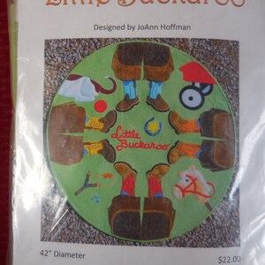 May include: A round, green, felt-like floor mat with a cowboy theme. The mat features a variety of cowboy-themed images, including boots, a horse, a lasso, and a wagon. The mat is 42 inches in diameter and is designed by JoAnn Hoffman.