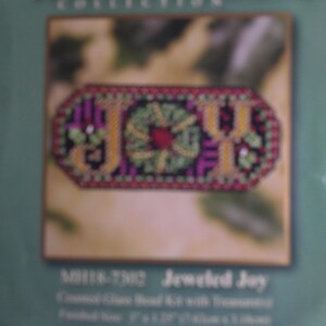 May include: A close-up of a craft kit with the words "JOY" in yellow, surrounded by a green wreath and pink and black accents. The kit is labeled "Winter Holiday Collection" and "Jeweled Joy". Finished size is 3" x 1.25" (7.6cm x 3.2cm).