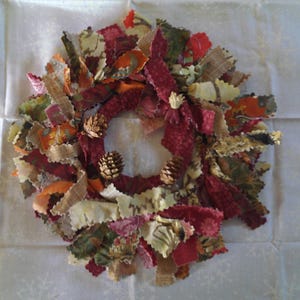 May include: A fall-themed wreath made of fabric scraps in shades of red, brown, cream, and green. The wreath is decorated with small pine cones.