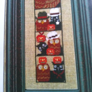 May include: A framed cross-stitch sampler on tan fabric featuring a vertical arrangement of four panels. Each panel depicts red owls, black cats, and orange jack-o'-lanterns in various hats and accessories, all enclosed in a dark green frame.