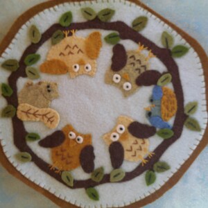 May include: A felt wall hanging with a brown border and a light blue center. The center features a tree branch with five owls and a squirrel, all made of felt in various shades of brown, beige, and blue.