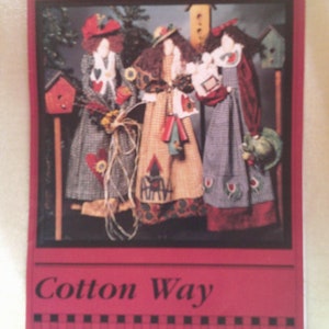 May include: Folkart Friends Cotton Way pattern featuring three dolls dressed in colorful patchwork dresses and hats. The dolls are standing in front of a wooden fence with birdhouses and flowers.