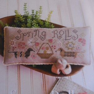 Puede incluir: Primer plano de una almohada bordada en rosa y marrón con el texto "Spring Rolls" en una fuente decorativa. La almohada está sentada en un cuenco de madera con plantas verdes.