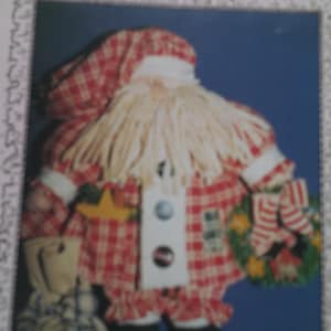 Peut inclure: Poupée artisanale du Père Noël avec une tenue à carreaux rouge et blanc, une barbe blanche et des bottes noires. La poupée tient une étoile et un petit sac. Une couronne verte avec un nœud rayé rouge et blanc est à droite.