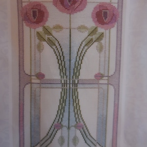 Può includere: Un ricamo a punto croce con tre papaveri rosa con foglie e steli verdi, all'interno di un disegno geometrico di linee blu, verdi e viola.