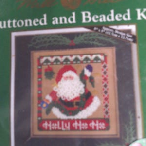 Può includere: Un kit per punto croce con un disegno di Babbo Natale. L'immagine mostra un ricamo incorniciato con Babbo Natale in abito rosso, barba bianca e le parole "Holly Ho Ho" in rosso. Il kit è etichettato "Buttoned and Beaded Kit".