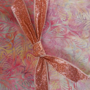Puede incluir: Una tela rosa y amarilla con un patrón floral está envuelta con una cinta de color oro rosa. La cinta está atada en un lazo en el centro de la tela.