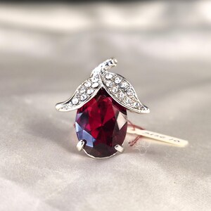 May include: A silver-toned ring featuring a large, faceted, oval-cut, deep red gemstone. The ring is accented with a leaf-shaped design encrusted with small, clear crystals. The ring is set against a soft, neutral background.