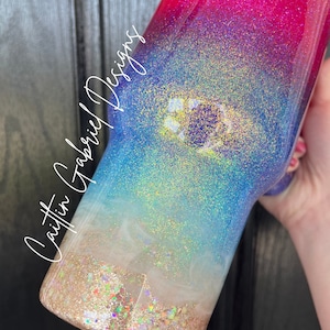 Puede incluir: Un vaso con purpurina con un dise&ntilde;o degradado, que va del rojo al rosa, azul y dorado. La secci&oacute;n inferior tiene un aspecto arenoso, como de playa, con purpurina iridiscente. El texto "Caitlin Gabriel Designs" est&aacute; escrito en el lateral.