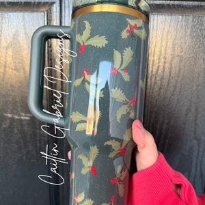 Op de afbeelding: Een donkerblauwe reismok met een handvat, met een hulst- en bessenpatroon. De mok heeft een gouden rand en een donkerblauwe deksel. De tekst "Caitlin Gabriel Designs" staat op de zijkant van de mok.