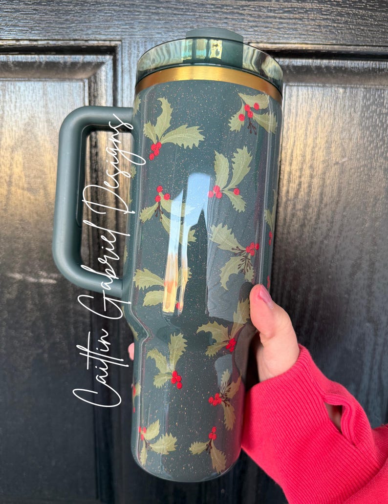 Op de afbeelding: Een donkerblauwe reismok met een handvat, met een hulst- en bessenpatroon. De mok heeft een gouden rand en een donkerblauwe deksel. De tekst "Caitlin Gabriel Designs" staat op de zijkant van de mok.