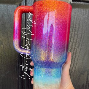 Puede incluir: Un vaso grande cubierto de purpurina con asa, que presenta un degradado de colores del rojo al azul, con un fondo arenoso similar a una playa. El vaso tiene una tapa blanca y el texto "Caitlin Gabriel Designs" en una fuente de escritura.