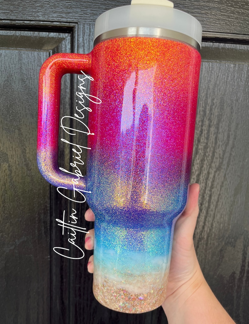 Puede incluir: Un vaso grande cubierto de purpurina con asa, que presenta un degradado de colores del rojo al azul, con un fondo arenoso similar a una playa. El vaso tiene una tapa blanca y el texto "Caitlin Gabriel Designs" en una fuente de escritura.