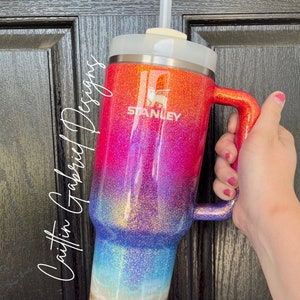 Puede incluir: Vaso Stanley brillante con un dise&ntilde;o degradado que va del naranja al rosa, azul y beige arenoso en la base. El vaso tiene una tapa blanca, un asa y una pajita transparente. El logotipo de Stanley es visible.