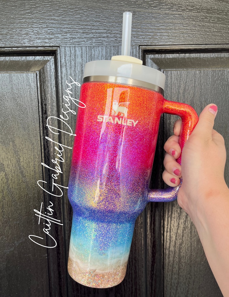 Puede incluir: Vaso Stanley brillante con un dise&ntilde;o degradado que va del naranja al rosa, azul y beige arenoso en la base. El vaso tiene una tapa blanca, un asa y una pajita transparente. El logotipo de Stanley es visible.