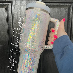 May include: A large, glitter-covered tumbler with a handle and a straw. The tumbler is white with iridescent glitter and a leopard print design. The text "Caitlin Gabriel Designs" is written on the side.