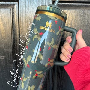 Op de afbeelding: Een donkerblauwe Stanley-beker met een gouden rand en handvat, met een hulst- en bessenpatroon. De beker heeft een rietje en het Stanley-logo in goud. De tekst "Caitlin Gabriel Designs" staat op de zijkant.