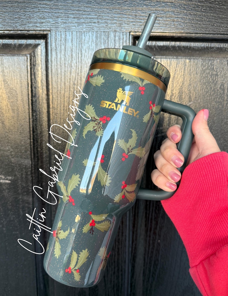Op de afbeelding: Een donkerblauwe Stanley-beker met een gouden rand en handvat, met een hulst- en bessenpatroon. De beker heeft een rietje en het Stanley-logo in goud. De tekst "Caitlin Gabriel Designs" staat op de zijkant.
