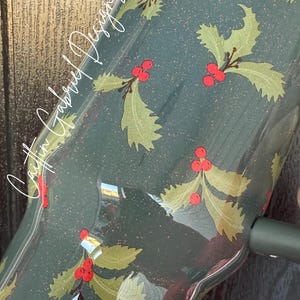 Op de afbeelding: Een donkergroene Stanley-beker met een feestelijk hulstbessenontwerp. De beker heeft een patroon van groene hulstbladeren en rode bessen, met de tekst "Caitlin Gabriel Designs" in witte letters. Het ontwerp is afgewerkt met glitter.