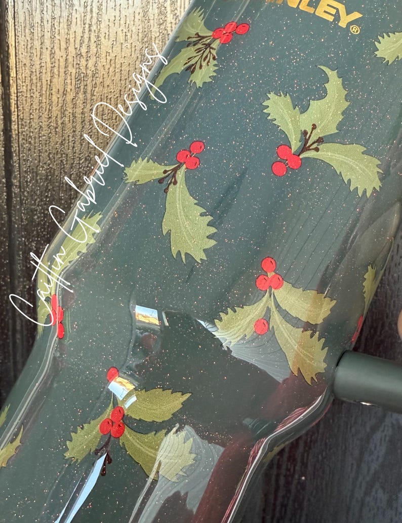 Op de afbeelding: Een donkergroene Stanley-beker met een feestelijk hulstbessenontwerp. De beker heeft een patroon van groene hulstbladeren en rode bessen, met de tekst "Caitlin Gabriel Designs" in witte letters. Het ontwerp is afgewerkt met glitter.