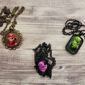 May include: Three necklaces with pendants featuring a fetus in a fetal position. The pendants are in different colors: red, green, and purple. The necklaces are on a wooden surface.