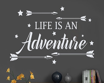 May include: White wall decal with the text "Life is an Adventure" and arrows and stars. The decal is on a grey wall above a wooden shelf with decorative items.