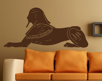 May include: A brown silhouette of an Egyptian sphinx with a human head and a lion's body. The sphinx is lying down with its head turned to the side. The silhouette is on a beige wall above a brown sofa with orange cushions.