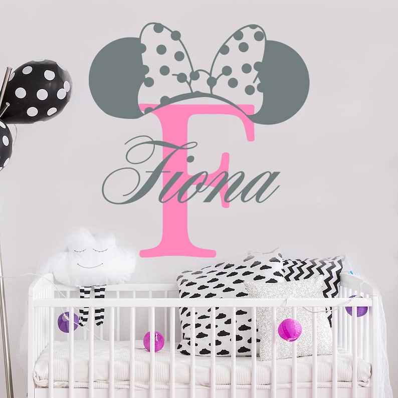 Puede incluir: Una cuna blanca con una calcoman&iacute;a de Minnie Mouse rosa y gris con la letra F y el nombre Fiona. La cuna tiene un colch&oacute;n blanco y una manta blanca. Hay almohadas con estampado blanco y negro en la cuna.