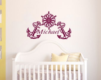May include: A white cot with a burgundy anchor and steering wheel wall decal with the name "Michael" written underneath.