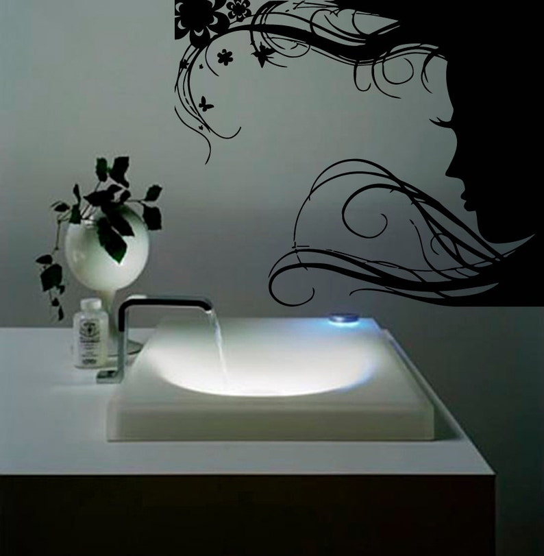 May include: A white ceramic sink with a chrome faucet and a flowing stream of water. The sink is illuminated from below, casting a soft glow. A black and white silhouette of a woman's face with flowing hair is on the wall behind the sink.
