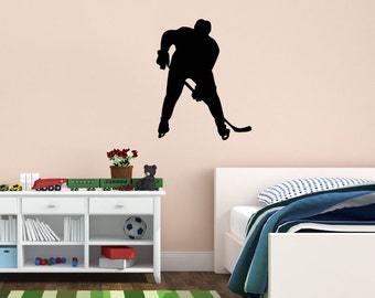 May include: Black silhouette of a hockey player with a stick, on a light pink wall, above a white bed with blue and white bedding, a white bedside table with a toy train, a stuffed animal, and a football, and a green and white striped rug.