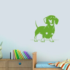 May include: Green silhouette of a dachshund dog standing on a white surface. The dog is facing forward with its ears perked up. The silhouette is against a light blue wall.