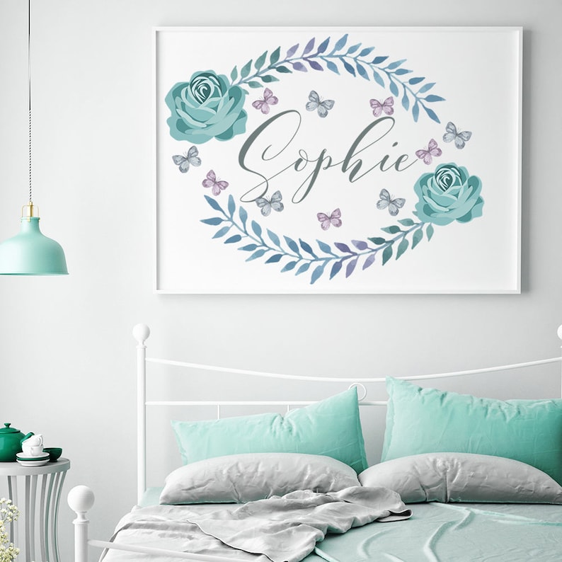 Puede incluir: Un cuadro enmarcado con el nombre "Sophie" en escritura elegante, rodeado por una corona de hojas azules, rosas turquesas y mariposas. La obra de arte se exhibe sobre una cama con almohadas turquesas y una colcha gris.