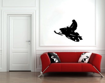 May include: A black silhouette of a person riding a snowmobile on a white wall. The snowmobile is facing the right side of the image. The person is wearing a helmet and a jacket. The snowmobile has a long track and a ski on the front. The snowmobile is on a white wall. There is a red sofa in the foreground.