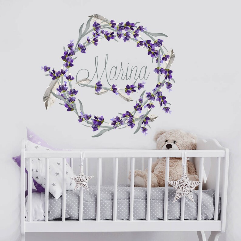 May include: A nursery wall decal featuring a lavender wreath with the name "Marina" in cursive script. The wreath is adorned with purple flowers and gray-green leaves. A white crib with star-shaped ornaments and a teddy bear is in the foreground.